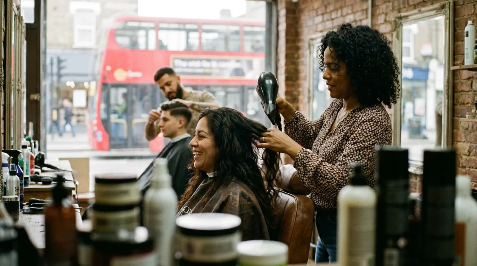 Estilista secando el pelo a una clienta en una peluquería latina de Londres, con el barbero al fondo