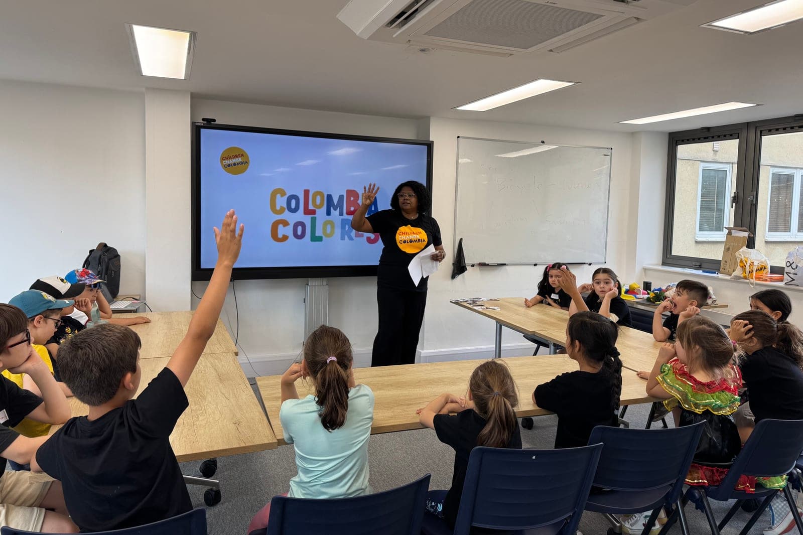 Una profesora de Colombia Colores, con la camiseta de Children Change Colombia, da la clase a un grupo de niños; uno levanta la mano. Detrás, una pantalla con el nombre del taller